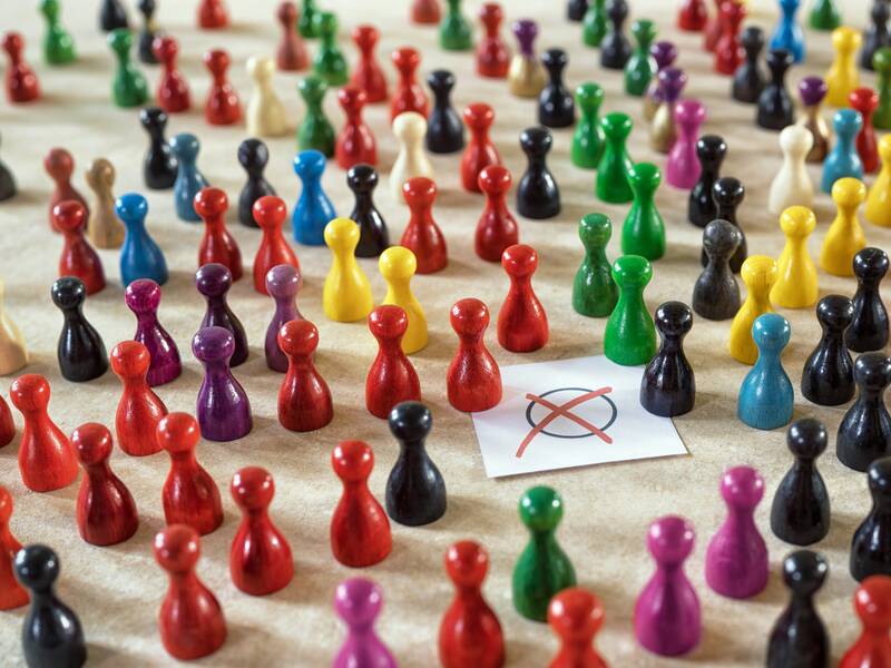 Ein Haufen bunter Spielfiguren steht auf einem Tisch, neben einem kleinen Blatt Papier mit einem roten Kreuz darauf.