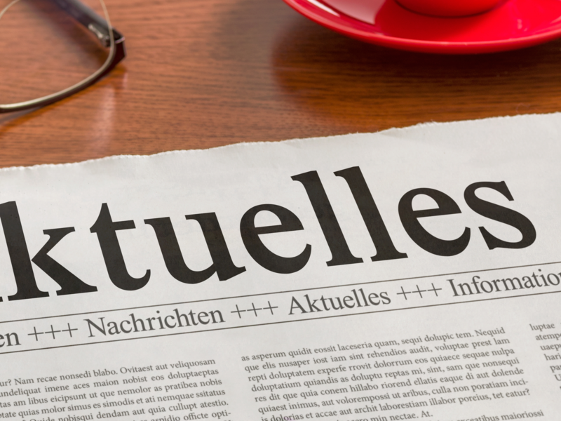 Eine Zeitung mit der Überschrift "Aktuelles" liegt auf einem Tisch neben einer Brille, einer Computertastatur und einem Stift.