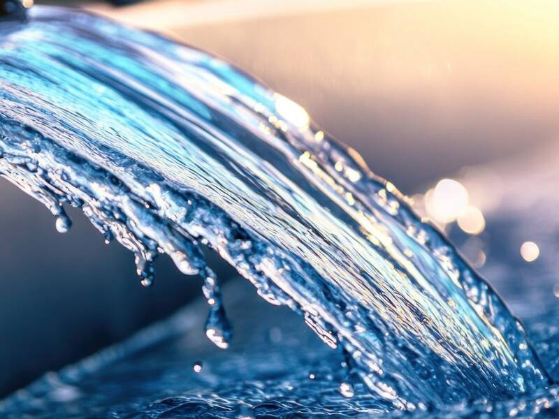Ein sanfter Wasserstrom fließt über die Kante eines schwarzen Behälters, schimmernd im Sonnenlicht.