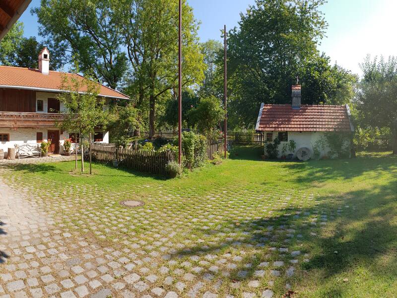 Ein ländlicher Hof mit einem traditionellen Gebäude und einem kleinen Nebengebäude, umgeben von Bäumen und einem gepflasterten Innenhof.