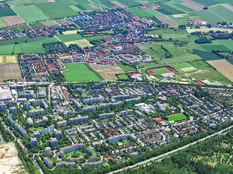 Das Bild zeigt eine Luftaufnahme einer weitläufigen Siedlung mit vielen Wohngebäuden, umgeben von landwirtschaftlichen Feldern und vereinzelten Wäldern.