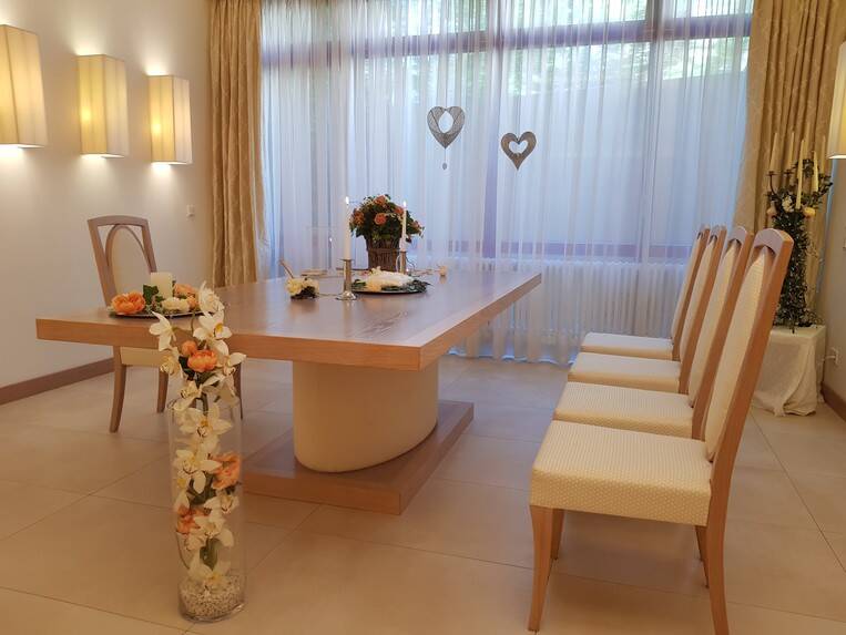 Blick in das Trauzimmer der Gemeinde. Ein stilvoll dekorierter Raum mit einem großen Tisch, darauf Blumen und Kerzen, umgeben von Stühlen, mit Vorhängen und herzförmigen Dekorationen im Hintergrund.