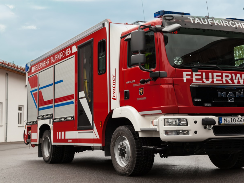 Das Bild zeigt ein rotes Feuerwehrfahrzeug mit der Aufschrift "Feuerwehr Taufkirchen" auf dem Parkplatz vor einem Gebäude.
