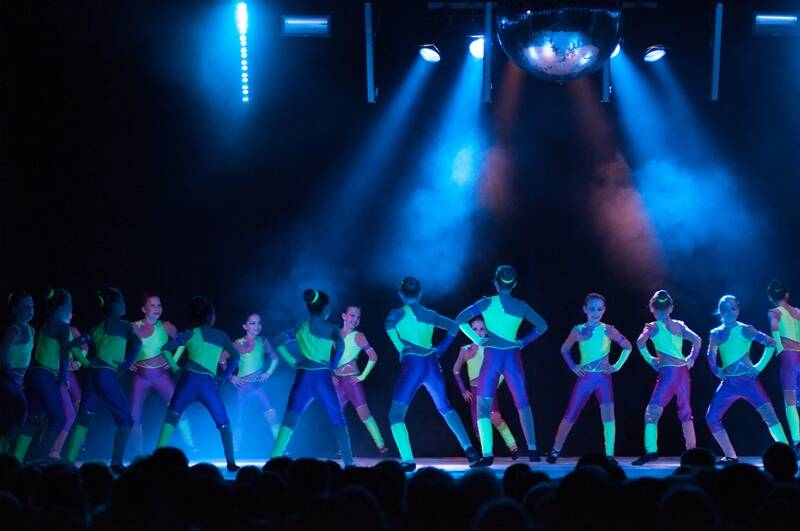 Eine Tanzgruppe mit Neon Kleidung steht auf der dunklen Bühne im blauen Scheinwerferlicht
