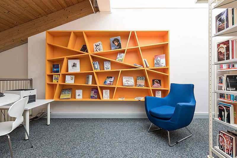 Ein oranges Bücherregal hängt gefüllt mit Büchern an einer weißen Wand mit einem blauen Stuhl davor