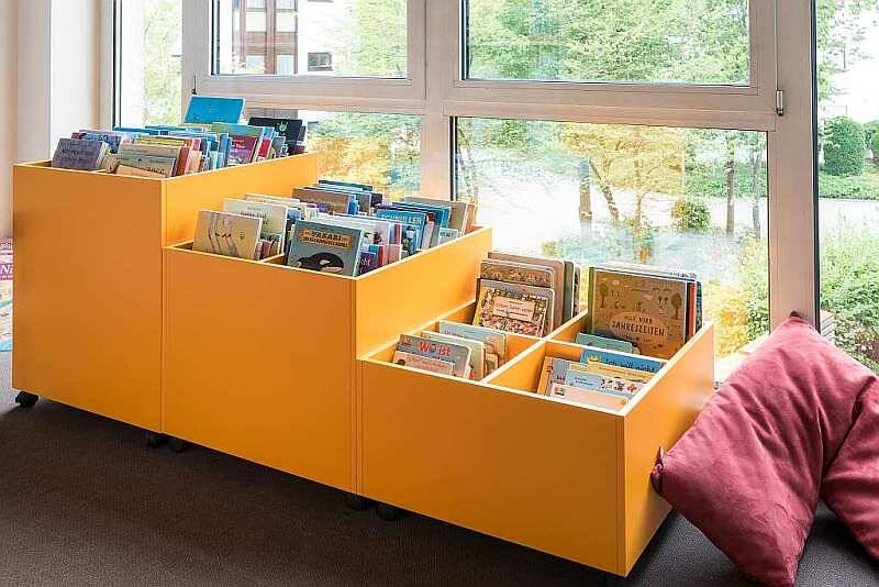 Ein oranger Büchertrog gefüllt mit Büchern steht vor einem Fenster