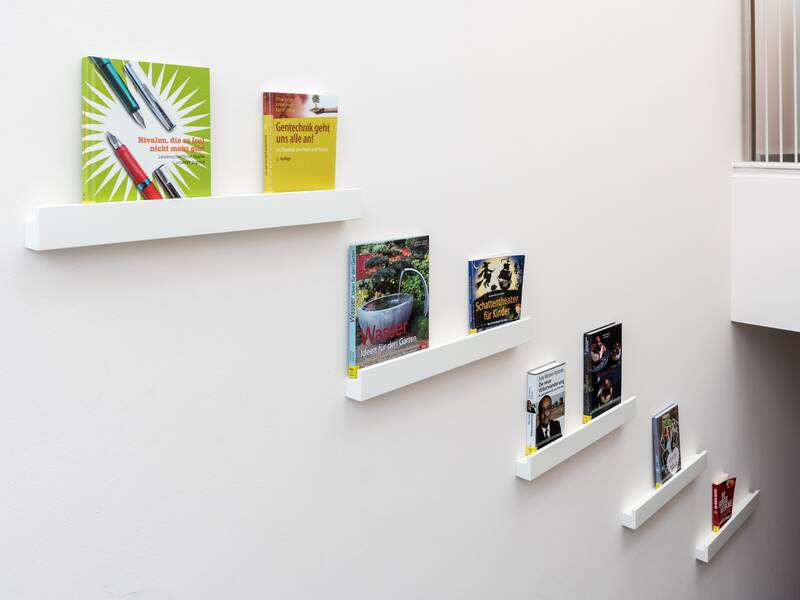 An einer weißen Wand hängen treppenförmig angeordnete Bücherregale wo jeweils zwei Buch mit bunten Titelseiten daruf steht