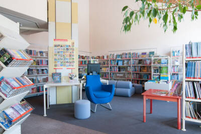 Ein Blauer Sessel steht umgeben von gefüllten Bücherregalen auf einem grauen Teppich mit einem roten Holztisch daneben