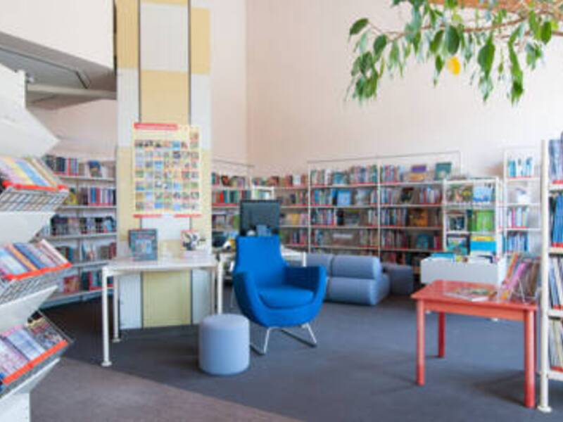 Ein Blauer Sessel steht umgeben von gefüllten Bücherregalen auf einem grauen Teppich mit einem roten Holztisch daneben