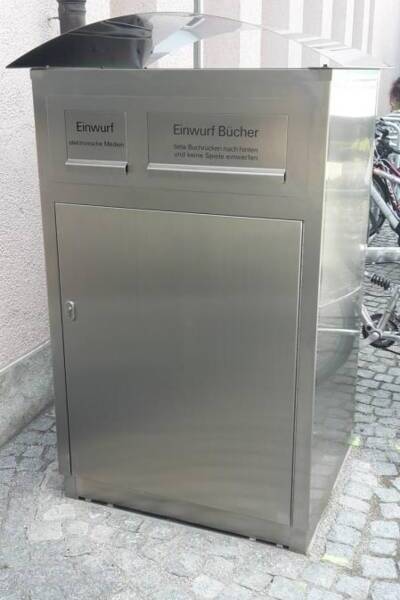 Eine silberne Medienrückgabebox mit zwei Fächern auf denen stehen die Worte Einwurf und Einwurf Bücher, steht auf einem gepflasterten Boden vor der Bücherei 