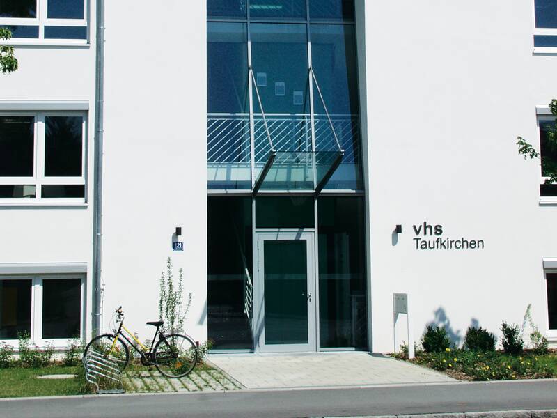 Ein weißes Haus mit einem Glaseingang in der Mitte, mit blauem Himmel und der Schrift VHS Taufkichen an der Wand neben dem Eingang, Davor steht ein dunkles Damenfahrrad