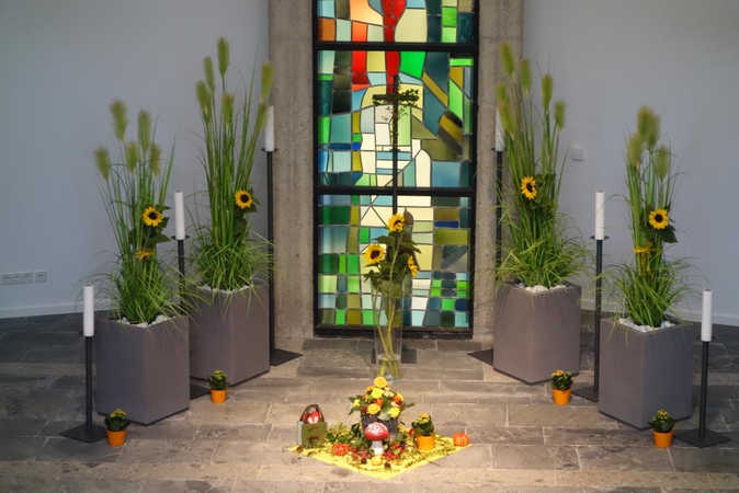 Buntes Mosaikfenster in der Kirche. Im Vordergrund stehen sechs weiße Kerzen und Stelen mit Pflanzen geschmückt auf dem Marmorboden