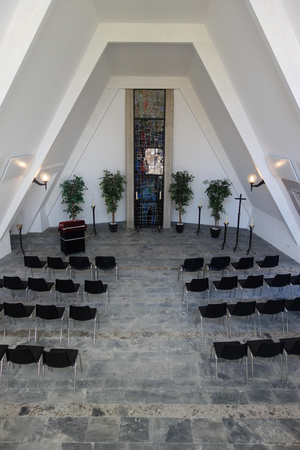 Schwarze Stühle stehen in der Kirche auf einem grauen Boden. Im Hintergrund befindet sich ein buntes Mosaikfenster vor dem rechts und links Pflanzen stehen und ein Rednerpult