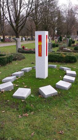 graue Marmor Stele mit orangem Bild, umgeben von liegenden Marmor Würfeln auf einer grünen Wiese. Im Hindergrund sind Gräber zu sehen