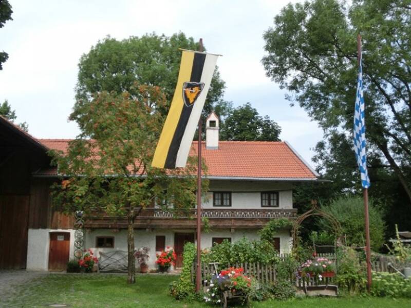 Ein weißes Haus  steht in einem üppigen, von Bäumen und Pflanzen umgebenen Garten mit einer Fahne davor, hinter einem Zaun mir bunten Blumen davor, unter einem strahlend blauen Himmel.