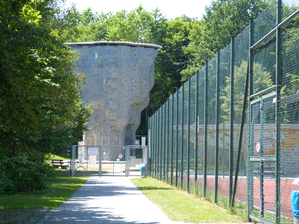 Ein Weg der zu einem Kletterfels führt mit einem hohen Zaun und grünen Bäumen daneben