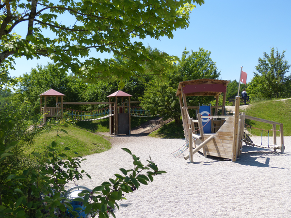 Ein Spielplatz mit Sand, einem Holzgerüst umgeben von Bäumen und blauem Himmel