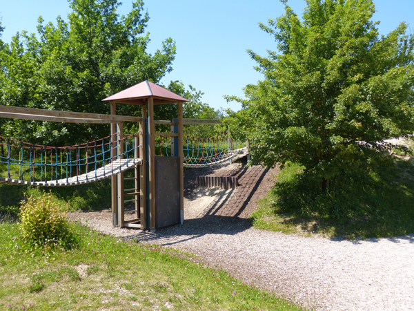 Ein Spielplatz mit Sand, einem Holzgerüst umgeben von Bäumen und blauem Himmel