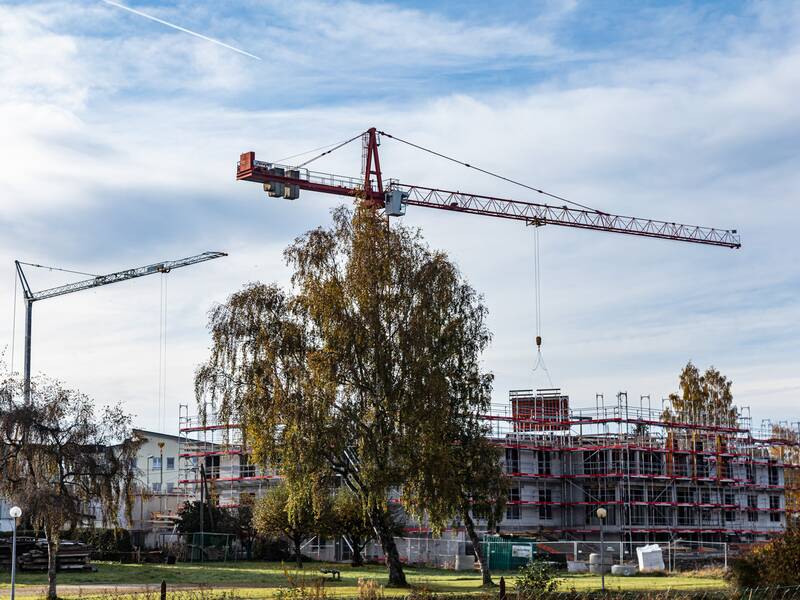 Eine Baustelle mit zwei großen Kränen und einem im Bau befindlichen Gebäude, umgeben von Bäumen, vor einem bewölkten Himmel.