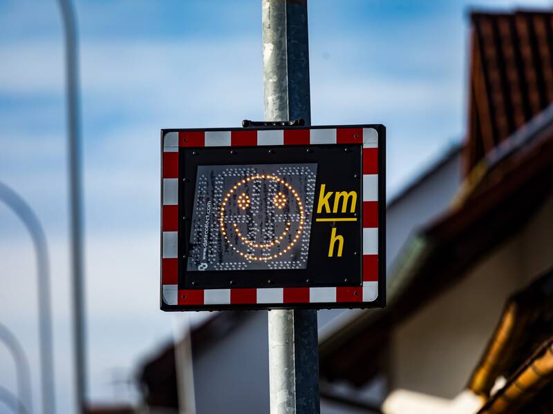 An einem Lichtmasten hängt ein rechteckiger, Geschwindigkeitsanzeige für den Straßenverkehr mit einem leuchtenden Smiley. daneben die Beschriftung km/h 
